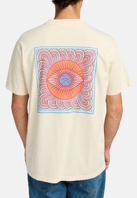 Camiseta de algodón color crema con un gran gráfico en la parte posterior que presenta un diseño central de un ojo naranja, rodeado de remolinos en tonos rosa, rojo y azul.