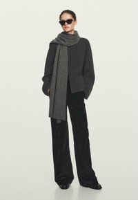 Giacca di lana grigia con chiusure a toggle, abbinata a una lunga sciarpa grigia. Pantaloni a zampa d'elefante in corduroy neri e occhiali da sole neri completano il look.