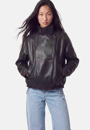 Femme portant une veste en cuir noire à col montant et un jean bleu clair, debout les mains dans les poches devant un fond uni.