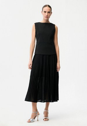 Frau steht vor weißem Hintergrund, trägt ein ärmelloses schwarzes Oberteil, einen plissierten schwarzen Midirock und silberne Sandalen mit Absatz und offenen Zehen.