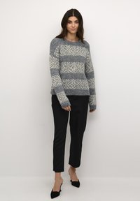 Pelēks adīts džemperis ar krēmkrāsas ģeometriskiem rakstiem, apaļa izgriezuma, garām piedurknēm un brīvu piegriezumu, saskaņots ar melnām piegriezuma biksēm un augstpapēžu kurpēm.