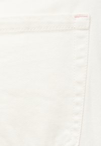 Κοντινή λήψη λευκού υφάσματος denim που δείχνει τσεπάκι σε σχήμα ορθογωνίου με ραφή και μια μικρή ροζ λεπτομέρεια στην πάνω γωνία.