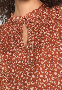 Blusa color ruggine con un motivo floreale, dotata di scollatura a forma di chiave e dettagli da annodare. Tessuto leggero con una superficie testurizzata.