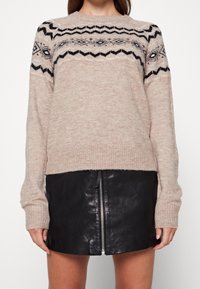 Maglione lavorato a maglia beige con motivi geometrici neri sul petto, scollo rotondo e polsini a costine, abbinato a una mini gonna in pelle nera.