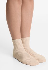 Calzini alla caviglia ribattuti beige con un motivo a maglia testurizzato, dotati di una parte superiore a risvolto e una cucitura liscia per le dita, appoggiati su gambe nude contro uno sfondo bianco.
