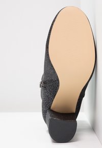 Svart texturerad sko med en slät sula, försedd med en sidoglidning och en låg chunky klack, som visar en subtil lyster i materialet.