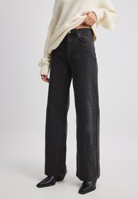 Jeans de pierna ancha en negro con una cintura alta, hechos de mezclilla. Presentan un estilo clásico de cinco bolsillos y un ligero desgaste, combinados con un suéter de punto color crema.