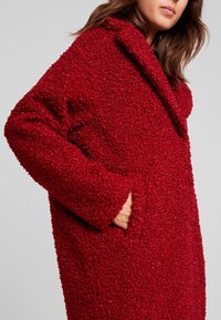 Červený huňatý kabát z tkaniny s texturovaným povrchem, volného střihu, širokým límcem a bočními kapsami, který nabízí pohodlný a teplý design.