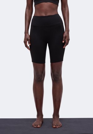 Kvinde stående barfodet på yogamåtte iført sorte højtaljede tætsiddende shorts og crop top mod en ensfarvet lys baggrund.