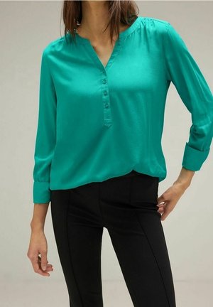 Femme portant un chemisier manches longues turquoise avec des boutons et un pantalon noir ajusté, posant devant un fond uni, montrant une partie de son torse.