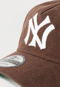 Καφέ καπέλο μπέιζμπολ με κεντημένο λευκό λογότυπο "NY", υφασμάτινη υφή και επίπεδο γείσο με κυκλικό ετικέτα μάρκας στο πλάι.