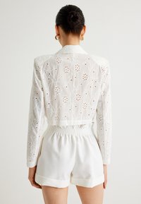 Vit broderad kavaj med blommönster, kombinerad med matchande vita shorts med resårmidja och upprullade fållar.