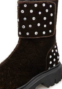 Stivale alla caviglia in suede marrone con texture a motivo, decorazioni in borchie e suola in gomma resistente. Risvoltato alla caviglia con pietre decorative.