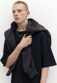 Fiatal férfi rövid vörös hajjal, fekete pólót visel, és sötét kapucnis pulóvert terít a vállára, oldalra nézve, fehér háttér előtt.