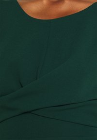 Zielona, teksturowana bluzka z okrągłym dekoltem i zwisającymi detalami z przodu, tworzącymi gładką, elegancką sylwetkę.