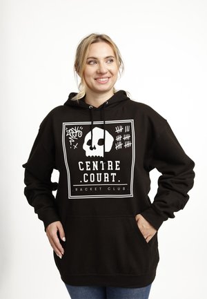 Kvinde iført en sort hættetrøje med en hvid kranium og teksten "Centre Court Racket Club", der smiler og kigger til siden.