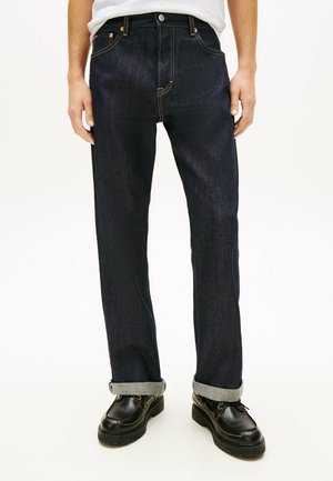 Mand iført mørkeblå denimjeans med opbukket kant og sorte læder snørestøvler, stående mod en ensfarvet lys baggrund.