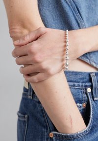 Bracelet en perles argentées attaché autour d'un poignet, avec des sphères réfléchissantes de tailles variées et une texture lisse. Chemise en denim et jeans visibles.