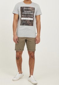 T-shirt gris avec un imprimé graphique comportant du texte, accompagné d'un short kaki et de baskets blanches. La tenue est décontractée et confortable.