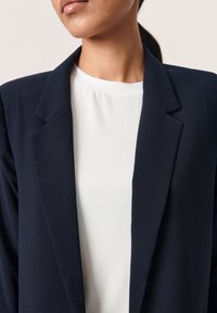 Námořnický blazer z hladkého materiálu s upraveným límcem, kombinovaný s bílým vrškem s kulatým výstřihem, který má volný střih a jemnou texturu.