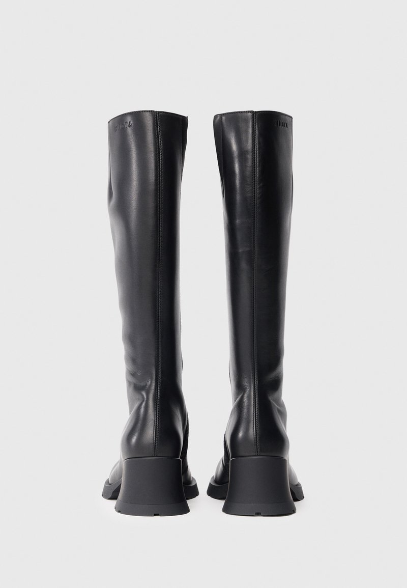 Knæhøje sorte læderstøvler med en glat tekstur, firkantet tå og chunky grå hæl. Har diskret branding på overdelen.
