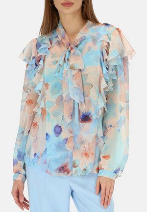 Kobieta w pastelowej, kwiatowej bluzce z falbanami na ramionach i dekoltem z wiązaniem, w połączeniu z jasnoniebieskimi spodniami.