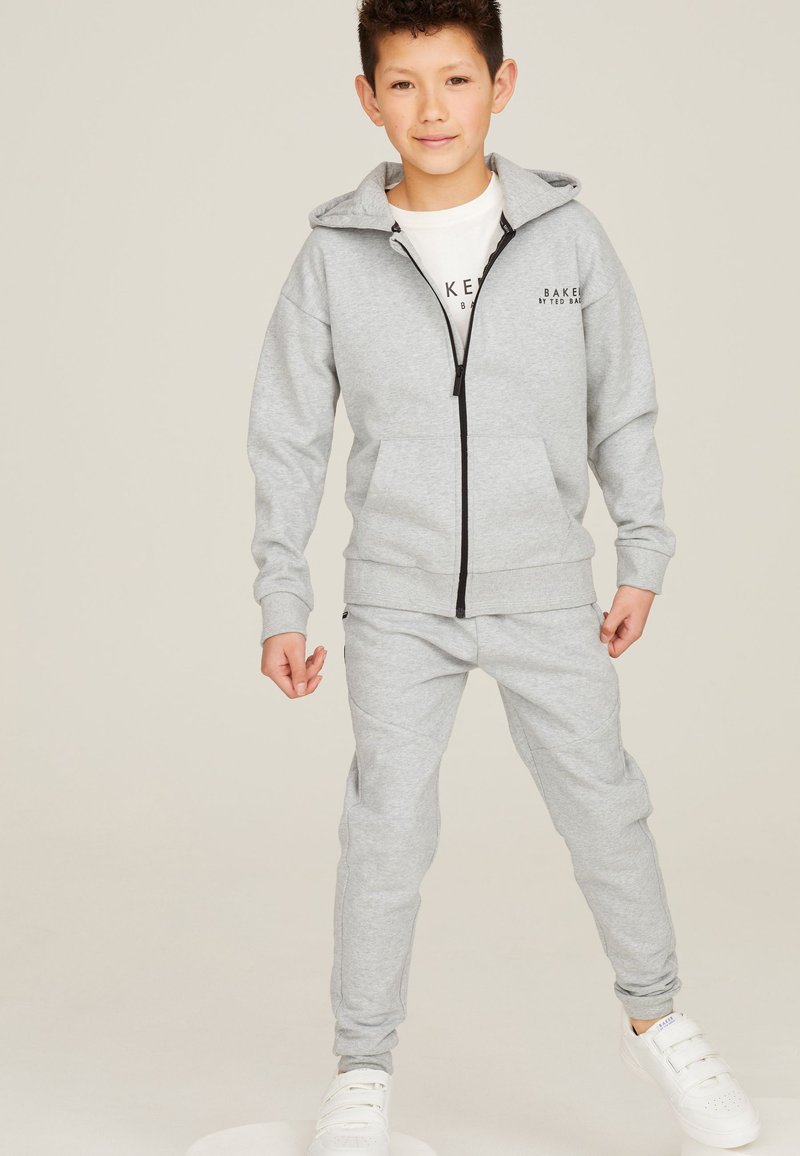 Garçon portant un sweat à capuche gris clair zippé et un pantalon de survêtement assorti avec des baskets blanches, debout devant un fond clair uni.
