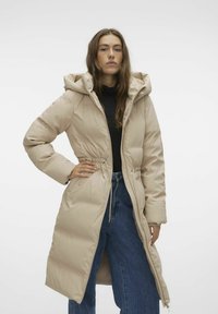 Manteau rembourré beige avec capuche amovible, taille cintrée, manches longues et fermeture éclair à l'avant, associé à un jean en denim bleu taille haute.