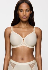 Beige rintaliivi, jossa on tekstuuriset kupit, pitsikoristeet olkaimissa ja koristeellinen etuosa. Sileä kangas, muotoiltu alaosa ja säädettävät olkaimet.