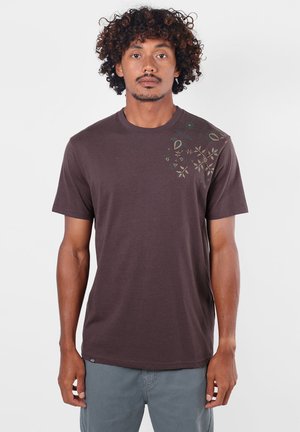 Homme aux cheveux bouclés portant un t-shirt marron foncé avec des motifs floraux clairs sur l'épaule, debout devant un fond blanc uni.
