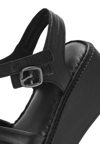 Sandale plateforme en cuir noir avec une sangle au niveau de la cheville dotée d'une boucle en argent, texture lisse et détails cousus le long du bord.