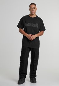 Tricou negru cu mâneci scurte și logo în relief, asortat cu pantaloni cargo negri cu mai multe buzunare și croială lejeră. Tenisi negri completează ținuta.