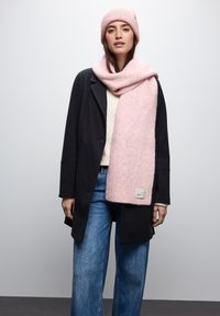 Una morbida sciarpa di maglia rosa pallido si adagia su un maglione crema, abbinata a un cappotto nero e a jeans a gamba larga blu chiaro, con un'etichetta sulla sciarpa.