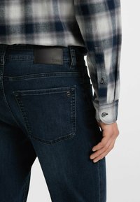 Jeans de mezclilla oscura con una textura suave, con bolsillos traseros y un parche de cuero. Combinados con una camisa de cuadros azul y blanca.