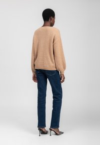 Beige, oversized sweater met een textuurafwerking, losse mouwen en een subtiele rugdetail. Gecombineerd met donkerblauwe straight-leg jeans en zwarte hakken.