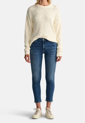Frau trägt einen cremefarbenen Pullover mit Zopfmuster, blaue Skinny-Jeans mit hoher Taille und cremefarbene Schnürsneaker, steht vor schlichtem Hintergrund.