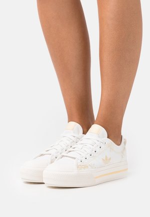 Weiße Leinensneaker mit goldenen Akzenten, runde Zehenpartie, flache Sohle und klassisches Schnürdesign. Texturierte Oberfläche und dezente Markenkennzeichnung.