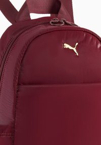 Zaino bordeaux in tessuto lucido; presenta una forma arrotondata, chiusura con zip e un accento dorato con logo. Superficie texturizzata con spallacci regolabili.