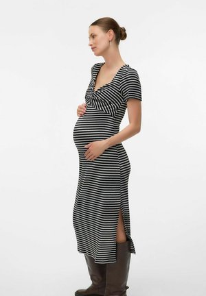Robe de maternité rayée noir et blanc avec des manches courtes, un col en V, des fentes sur les côtés et une coupe ajustée, portée avec des bottes marron montant jusqu'aux genoux.