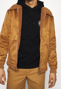 Veste marron en suédine synthétique avec col et poignets en peau lainée, comprenant un sweat à capuche noir en dessous et un pantalon marron assorti.