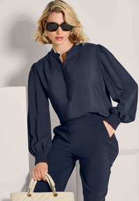 Frau mit kurzen blonden Haaren, die schwarze Sonnenbrille, eine marineblaue Bluse mit Puffärmeln und eine marineblaue Hose trägt und eine cremefarbene, gesteppten Handtasche hält.