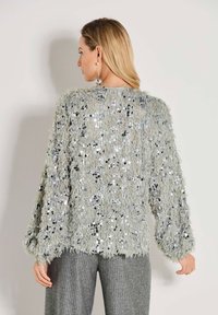 Eine Frau mit langen blonden Haaren trägt einen hellgrauen, kuscheligen Pullover mit silbernen Pailletten und grauen Nadelstreifen-Hosen und steht an einer hellen Wand.