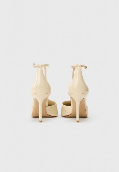Escarpins en cuir verni beige avec un bout pointu, une fine bride de cheville et un talon aiguille élancé. Texture lisse et détails minimaux.