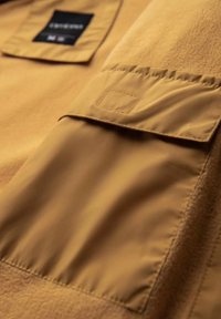 Brunt tyg med en klaffficka med rektangulär design. Texturen är slät, med kontrasterande sömmar och en subtil glans.