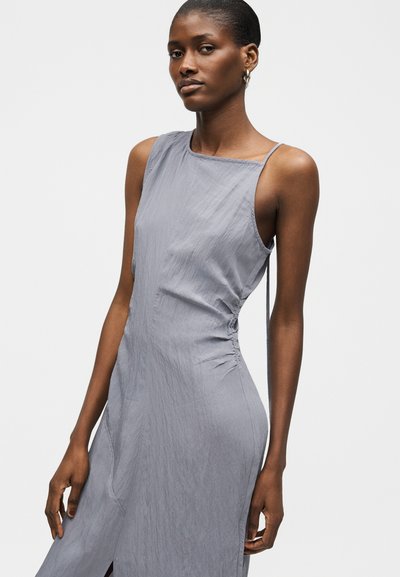 Νεαρή γυναίκα με κοντά μαλλιά που φοράει αμάνικο, ανοιχτό γκρι στενό φόρεμα με ρεντζετ side και σκίσιμο, στέκεται μπροστά σε ένα λευκό φόντο.