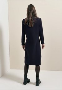 Robe en maille bleu marine, longueur genou, manches longues, tissu texturé, portée avec des bottines noires et des collants opaques, vue de dos.