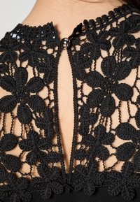 Schwarzer Blumen-Spitzenstoff mit durchbrochenem Detail, das mit einem kleinen glänzenden Knopf an der nackten Haut am Rückenausschnitt eines Kleidungsstücks befestigt ist.