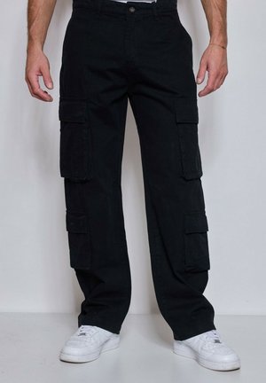 Homme portant un pantalon cargo noir avec de grandes poches latérales et des baskets blanches, debout contre un mur uni.