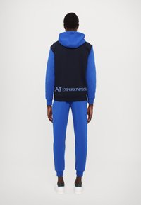 Completo tuta in blu navy e blu brillante con felpa con cappuccio e pantaloni coordinati, con dettaglio del logo in vita e nei polsini.