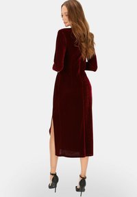 Bordowa aksamitna sukienka z długimi rękawami i bocznym rozcięciem, o dopasowanym kroju i gładkiej fakturze. Nosi się ją z czarnymi sandałami na obcasie.